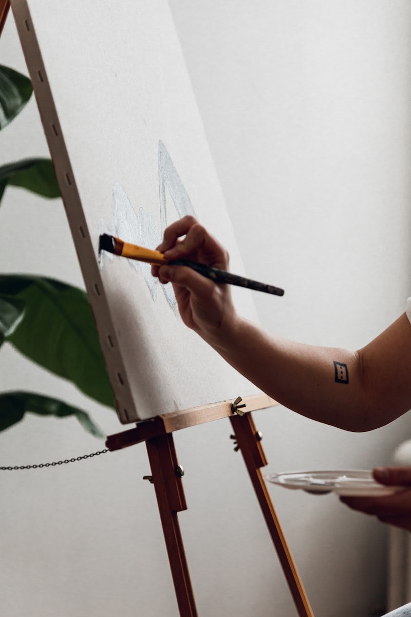 A hand loading a flat brush against a stretched canvas on an easel.
